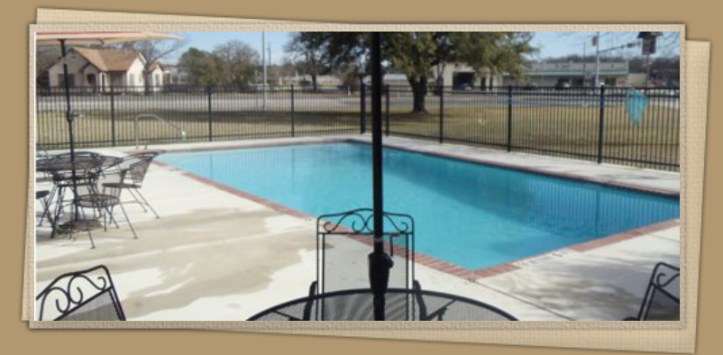 A fenced outdoor swimming pool at a Kingsland Texas Hotel, with patio tables and chairs around it, surrounded by trees, grass, and buildings in the background under a clear sky.